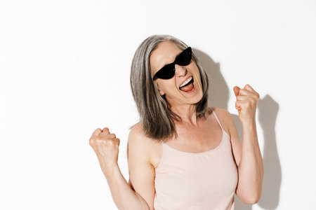 Mature woman in sunglasses making winner gesture while screaming isolated over white wallの写真素材