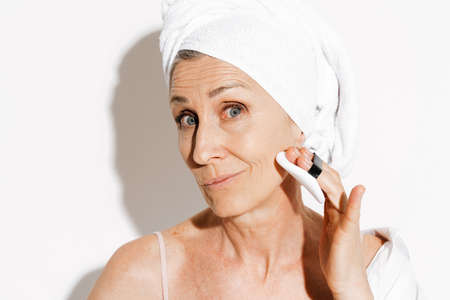 Senior woman wrapped in towel using powder sponge while doing makeup isolated over white wallの写真素材