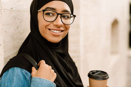 Close up of a smiling young muslim woman in hijab standing on a street using mobile phone drinking coffeeの写真素材