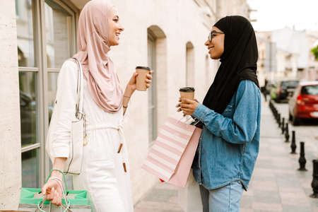 Two smiling mid aged islamic women in headscarves walking outside carrying shopping bags talking holding takeaway coffee cupsの写真素材