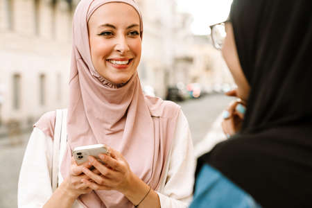 Multiracial muslim women wearing hijab talking while using mobile phone at city streetの写真素材