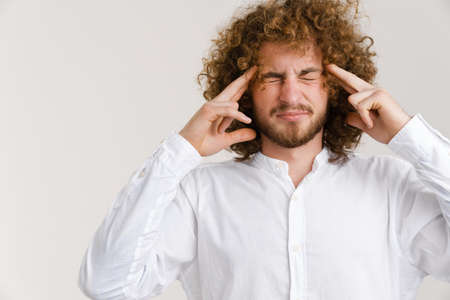 Young curly man with headache rubbing his temple and grimacing isolated over white backgroundの写真素材