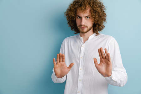 Young curly man with ginger hair showing stop gesture at camera isolated over blue backgroundの写真素材