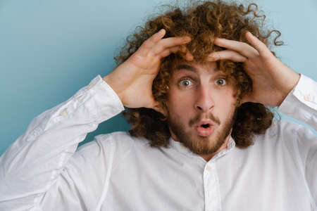Young man with ginger curly hair expressing surprise at camera isolated over blue wallの写真素材