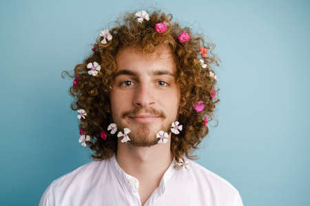 Close up portrait of a smiling young white man in shirt with flowers in his curly hair and beard standing over blue backgroundの写真素材