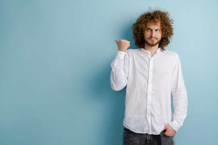 Young curly man with ginger hair frowning and pointing finger aside isolated over blue backgroundの写真素材