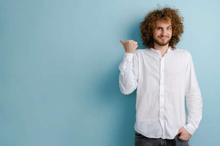 Young curly man with ginger hair frowning and pointing finger aside isolated over blue backgroundの写真素材