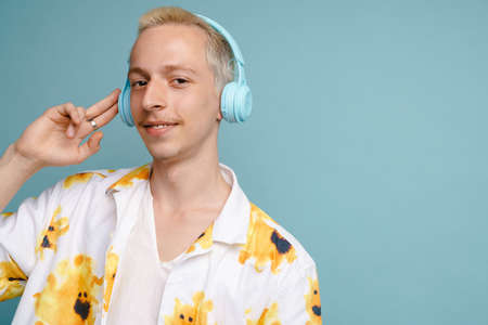 Young blonde man in shirt listening music with headphones isolated over blue background