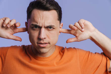Young brunette man frowning and plugging his ears isolated over purple background