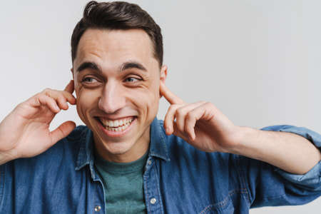 Young brunette man smiling and plugging his ears isolated over white background