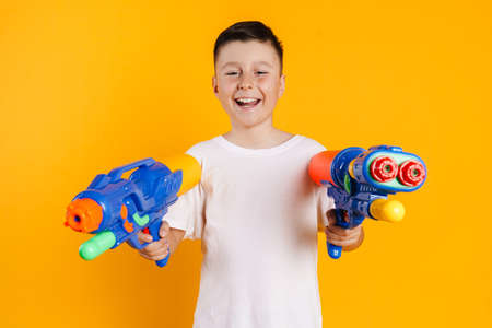 Happy smiling preteen boy in t-shirt standing over yellow background playing with watergun