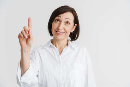 European mature woman smiling and pointing finger upward isolated over white wall