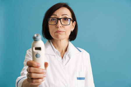 European mature doctor in eyeglasses while posing with thermometer isolated over blue wallの写真素材