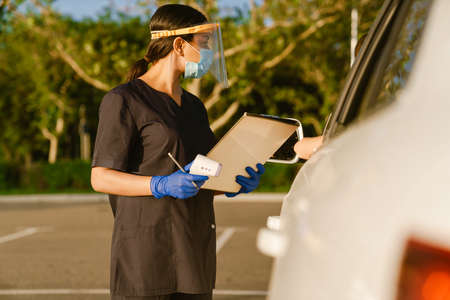 Black health worker in face mask and gloves writing down notes while standing by car outdoors