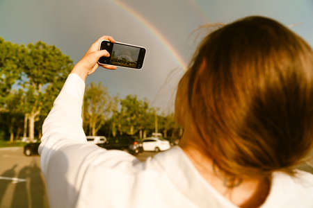 Young white woman taking photo of rainbow on cellphone at parking outdoors