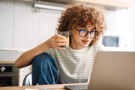 European curly woman using laptop and earphones while drinking juice at home kitchenの写真素材
