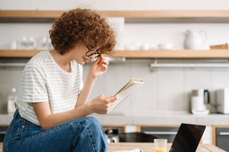 Young curly woman reading her notes while sitting on table at kitchenの写真素材
