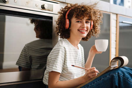 Smiling curly woman in headphones drinking coffee and drawing while sitting on floor at kitchen