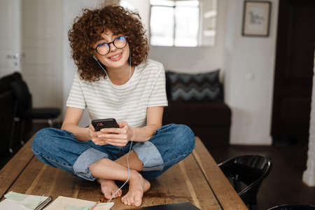Smiling young woman on the phone in the kitchen, sitting with earphones on table