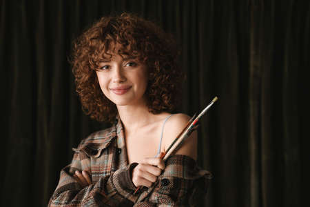 Smiling young redhead woman in plaid shirt standing in studio posing with paintbrush