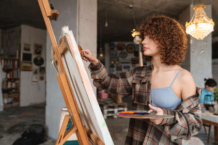 Smiling young white redhead woman artist painting standing in front of canvas in studio workshop