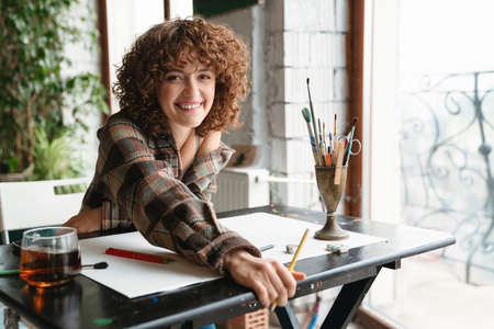 Smiling young redhead woman artist drawing with pencil sitting at the desk with cup of tea in the...