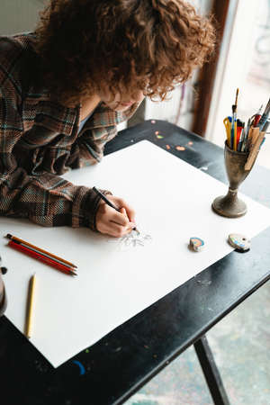 Young redhead woman artist drawing with pencil sitting at the desk with cup of tea in the studio workshopの写真素材