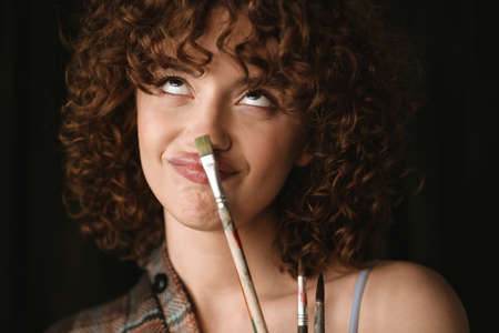 Smiling young redhead woman in plaid shirt standing in studio posing with paintbrush grimacing