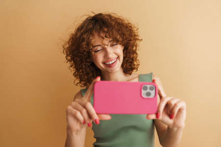 Young ginger woman smiling while taking photo on mobile phone isolated over beige background