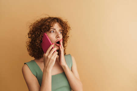 Excited shocked young casual woman with curly hair talking on mobile phone standing over beige...