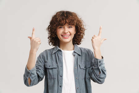 Young ginger woman in shirt smiling and pointing fingers upward isolated over white background