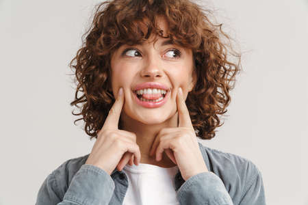 Ginger joyful woman showing her tongue and pointing fingers at her cheeks isolated over white...