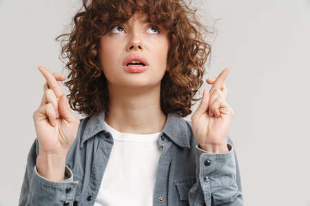 Young ginger unhappy woman holding fingers crossed for good luck isolated over white background
