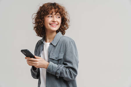 Young ginger joyful woman smiling while using mobile phone isolated over white background