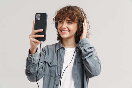 Young ginger joyful woman listening music with headphones and cellphone isolated over white...