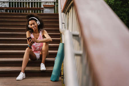 Young black sportswoman in headphones using cellphone while sitting on stairs outdoorsの写真素材