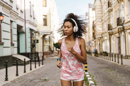 Young black woman listening music with headphones while running on city street outdoorsの写真素材