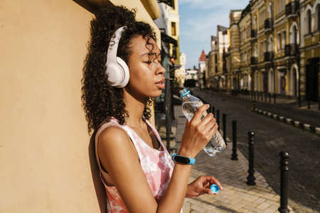 Young black woman in headphones drinking water during workout on city street outdoorsの写真素材
