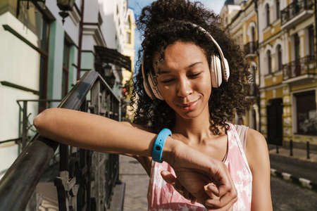 Young black woman in headphones using smartwatch during workout on city street outdoors