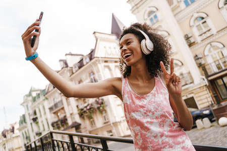 Black woman in headphones gesturing while taking selfie on cellphone at city street outdoorsの写真素材