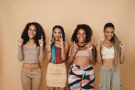 Young black four women laughing while pointing fingers upward together isolated over beige backgroundの写真素材