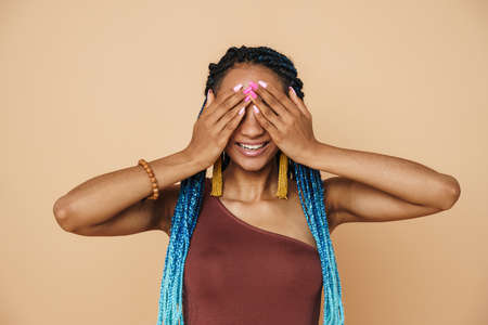 Black woman with blue afro pigtails smiling while covering her eyes isolated over beige backgroundの写真素材