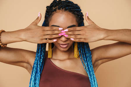 Young black woman with blue afro pigtails covering her eyes isolated over beige background