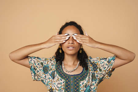 Young black woman wearing dress covering her eyes isolated over beige backgroundの写真素材