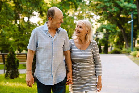 White senior couple holding hands and smiling while walking in summer parkの写真素材
