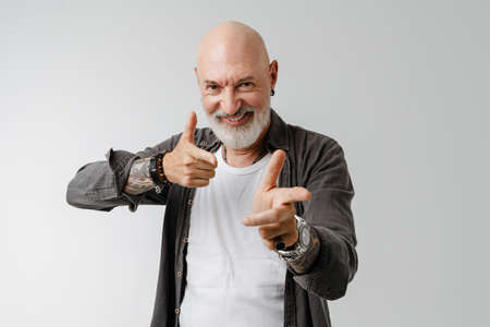 Bald european man with beard laughing while pointing fingers at camera isolated over white...