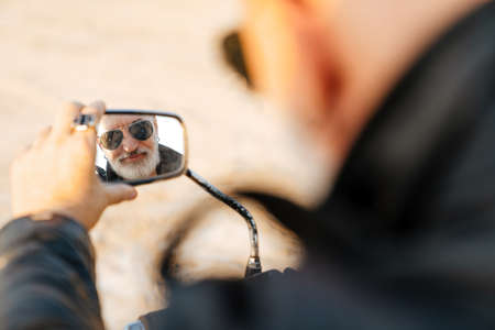 Mature bearded man looking in rear-view mirror on motorbike outdoors