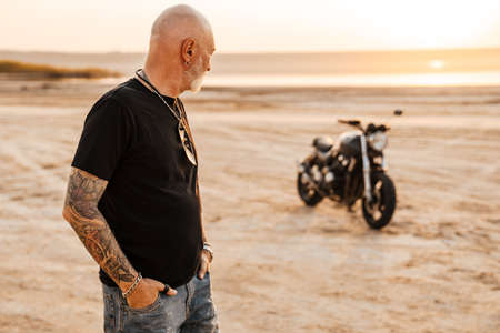 Bold senior man wearing t-shirt posing with motorcycle outdoors on summer dayの写真素材