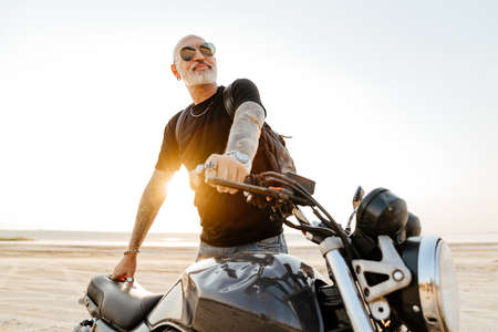 Bold senior man in sunglasses posing with motorcycle outdoors on summer day