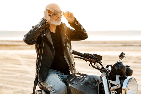Bold senior man wearing leather jacket posing on motorcycle outdoors on summer dayの写真素材
